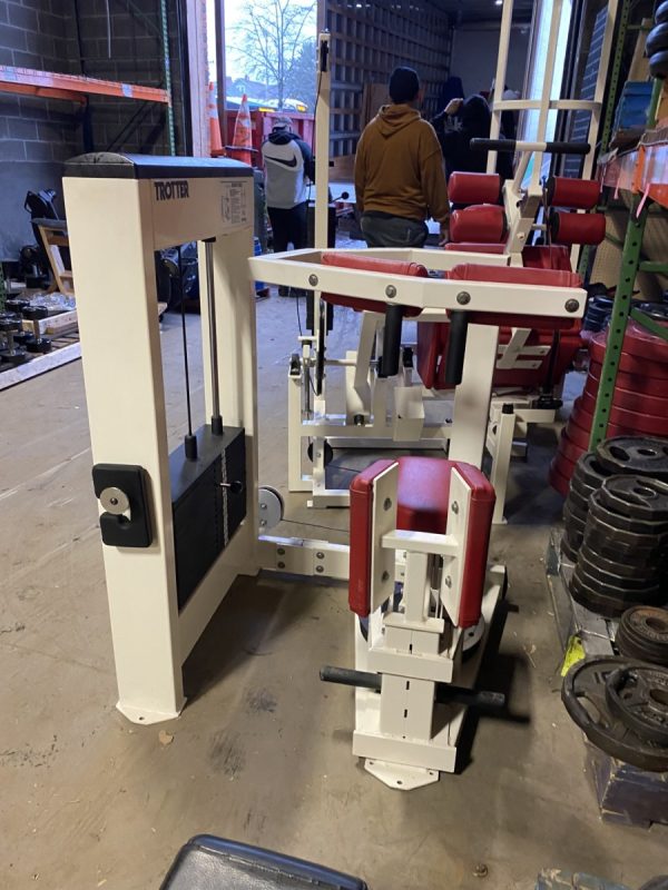 A Trotter Galileo Rotary Torso - As Is Functional exercise machine is positioned in a storage area, surrounded by various weights and equipment, with people visible in the background.