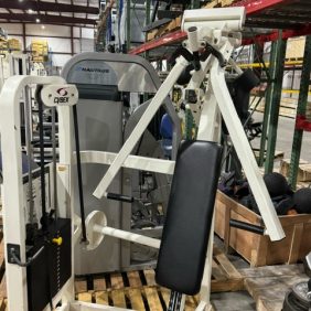 The gym equipment, such as the Cybex VR2 Converging Chest Press - As Is Functional, is placed on a wooden pallet inside a warehouse. In the background, shelving holds various functional items alongside a Nautilus machine.