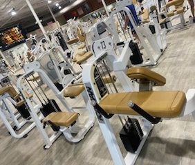 A gym interior showcasing the Icarian Package - Serviced, with rows of beige and white weight machines on a light-colored floor, meticulously maintained under bright overhead lighting in our fully serviced facility.