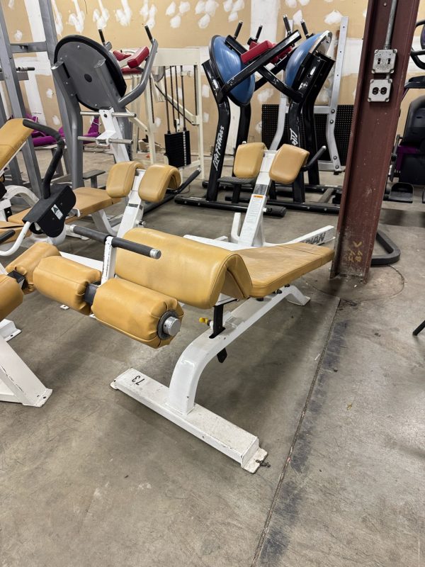 A gym leg extension and curl machine with tan upholstery and white frame is positioned on a concrete floor. Other gym equipment is visible in the background.