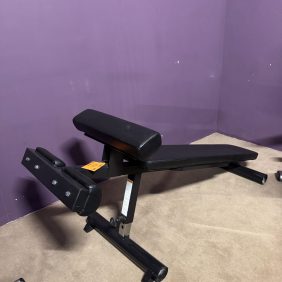 The Body Core Incline Bench - As Is Functional is designed for core workouts with black padding. It rests on a carpeted floor against a purple wall, offering adjustable incline capabilities for varied exercises.