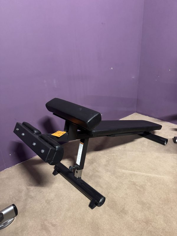 The Body Core Incline Bench - As Is Functional is designed for core workouts with black padding. It rests on a carpeted floor against a purple wall, offering adjustable incline capabilities for varied exercises.