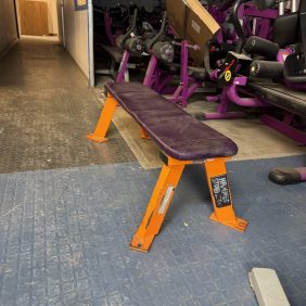 A gym showcases a purple and orange Hammer Strength Flat Bench - As Is Functional on a textured blue floor, with various exercise equipment like stationary bikes surrounding it.