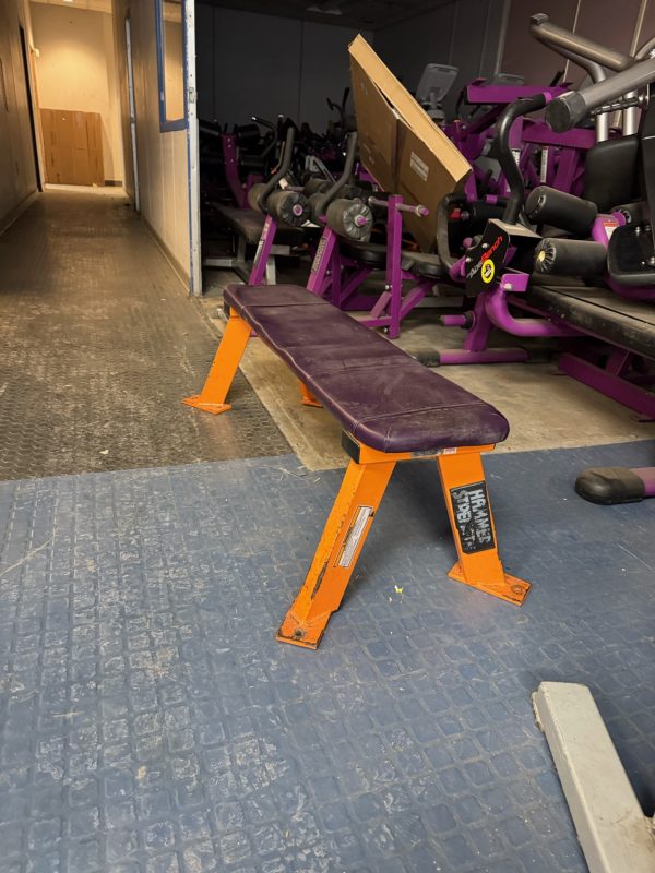 A gym showcases a purple and orange Hammer Strength Flat Bench - As Is Functional on a textured blue floor, with various exercise equipment like stationary bikes surrounding it.