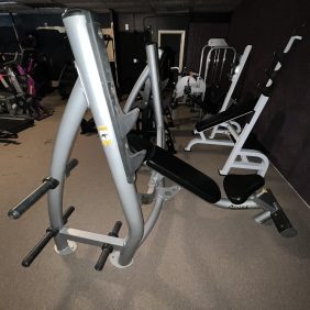 In a dim gym among the equipment, a remanufactured weight bench with padded seat and backrest stands next to the versatile Precor Stretch Machine - Remanufactured, inviting fitness enthusiasts to enjoy a comprehensive workout experience.