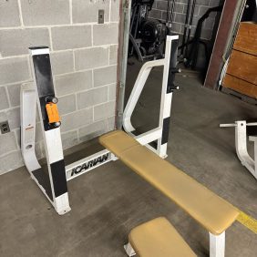 The True XFW 8200 Olympic 3 Way - New complements the beige padding and white frame of the old bench press, standing out against the gym's bare gray walls.