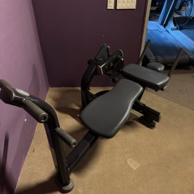 A sleek Life Fitness Signature Ab Crunch in black ($1995) sits in the corner against a vibrant purple wall, featuring cushioned supports and sturdy handles. Nearby, light switches are conveniently accessible.