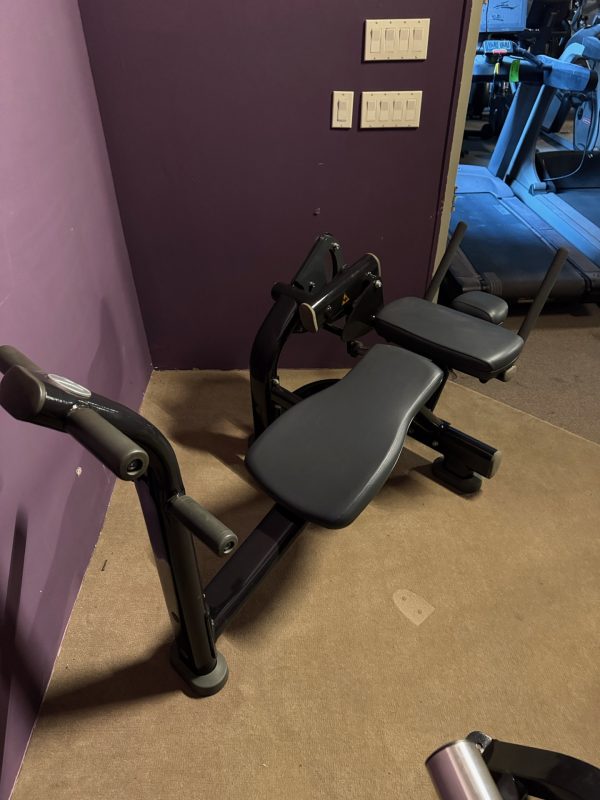 A sleek Life Fitness Signature Ab Crunch in black ($1995) sits in the corner against a vibrant purple wall, featuring cushioned supports and sturdy handles. Nearby, light switches are conveniently accessible.