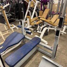 The cluttered room displays various fitness equipment, such as benches and weight machines, highlighted by the Icarian Glute Isolator ($995), alongside some pieces with blue and beige padding.