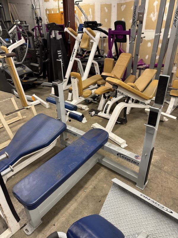 The cluttered room displays various fitness equipment, such as benches and weight machines, highlighted by the Icarian Glute Isolator ($995), alongside some pieces with blue and beige padding.