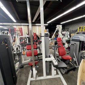 A gym features various weight machines, including a remanufactured Maxicam seated row with red padded seats and metal frames, along with a remanufactured Precor Stretch Machine.