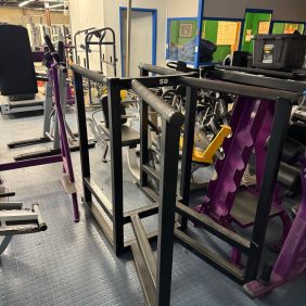 A gym with a versatile setup includes the Miscellaneous Dip Station Dual - As Is Functional and weight racks, all on a blue tiled floor.