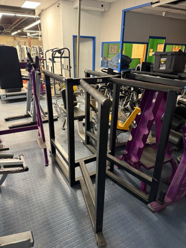 A gym with a versatile setup includes the Miscellaneous Dip Station Dual - As Is Functional and weight racks, all on a blue tiled floor.