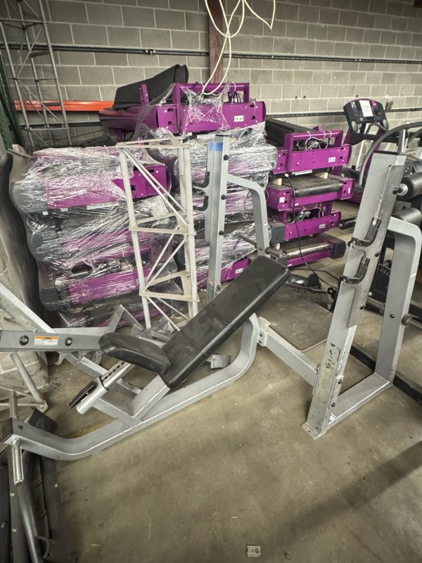 Gym equipment and machines stacked in a storage area with a concrete wall background.