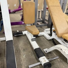 The Precor Utility Bench - As Is Functional, featuring tan cushions and a white metal frame, is ideal for seated exercises. Other fitness equipment surrounds it, with a partially painted wall in the background enhancing the gym's dynamic atmosphere.