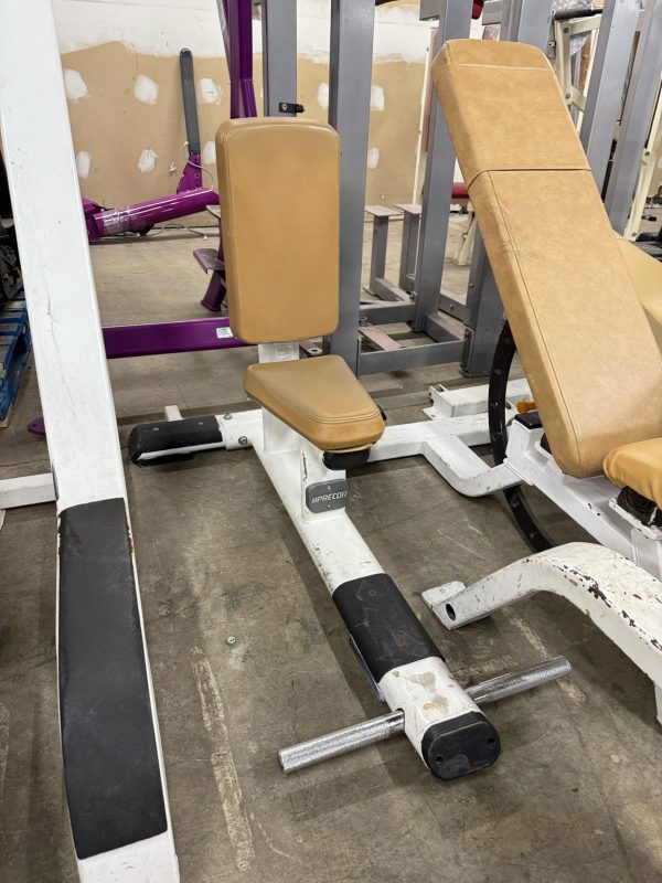 The Precor Utility Bench - As Is Functional, featuring tan cushions and a white metal frame, is ideal for seated exercises. Other fitness equipment surrounds it, with a partially painted wall in the background enhancing the gym's dynamic atmosphere.